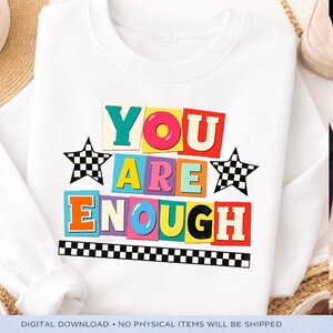 Pode incluir: Camisola branca com a frase "YOU ARE ENOUGH" em letras coloridas. O design inclui estrelas quadriculadas em preto e branco e uma borda quadriculada. A camisola é um download digital.