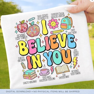 May include: White t-shirt with a colorful graphic design. The design includes the words "I Believe in You" in large, bubble letters, surrounded by various illustrations like an apple, disco ball, and a flower. Additional text includes motivational phrases.