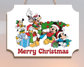 Könnte beinhalten: Ein weißes Holzschild mit dekorativem Rand, das ein Cartoon-Bild von Micky Maus, Donald Duck, Goofy und Pluto zeigt, die Weihnachten feiern. Auf dem Schild steht in roter Schrift "Merry Christmas". Zum Aufhängen ist eine Juteschnur befestigt.