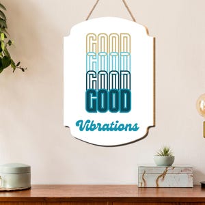 May include: A white wooden sign with a decorative shape, hanging by a rope. The sign features the word "GOOD" stacked four times in different shades of blue and gold, with "Vibrations" written below in teal.