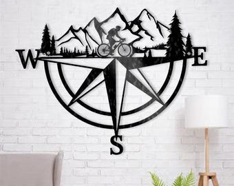 May include: Black metal wall art featuring a compass design with a cyclist riding through mountains. The compass points to the cardinal directions, with the letters W, E, and S visible. The artwork is mounted on a white brick wall.