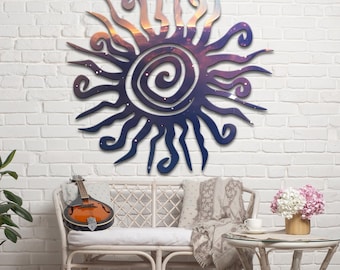 May include: A large, dark blue metal sunburst wall art with a spiral centre and stylized rays. The centre and rays feature a gradient of colours, including orange, purple, and blue. The sun is mounted on a white brick wall.