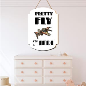 Könnte beinhalten: Weißes Holzschild mit einer Seilaufhängung. Das Schild zeigt den Text "PRETTY FLY FOR A JEDI" in Schwarz, mit einer Illustration eines Sternenjägers. Das Schild hängt über einer weißen Kommode.