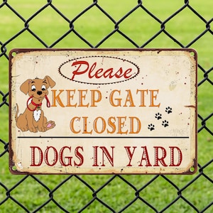 Op de afbeelding: Een rechthoekig metalen bord met een verweerde afwerking, met een cartoonhond en de tekst "Please Keep Gate Closed Dogs in Yard". Het bord wordt omlijst door een zwart gaashek tegen een groene grasachtergrond.