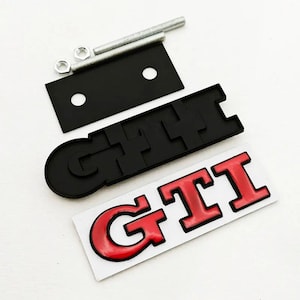 May include: A selection of car emblems and fixings. Features a black GTI emblem, a red and white GTI sticker, a black mounting plate, nuts, and a threaded rod. The GTI emblems are in a bold, blocky font.