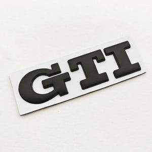May include: Black GTI badge on a white background. The letters are thick and raised, giving a 3D appearance. The badge is rectangular and probably for automotive use.