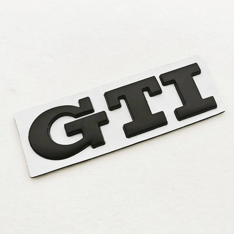 May include: Black GTI badge on a white background. The letters are thick and raised, giving a 3D appearance. The badge is rectangular and probably for automotive use.