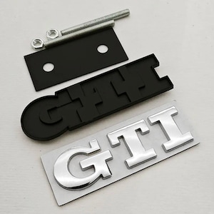 May include: A black and silver GTI emblem set, comprising a black base, silver letters, mounting hardware, and a black mounting plate. The GTI letters are in a bold, chrome-like font. Designed for automotive applications.