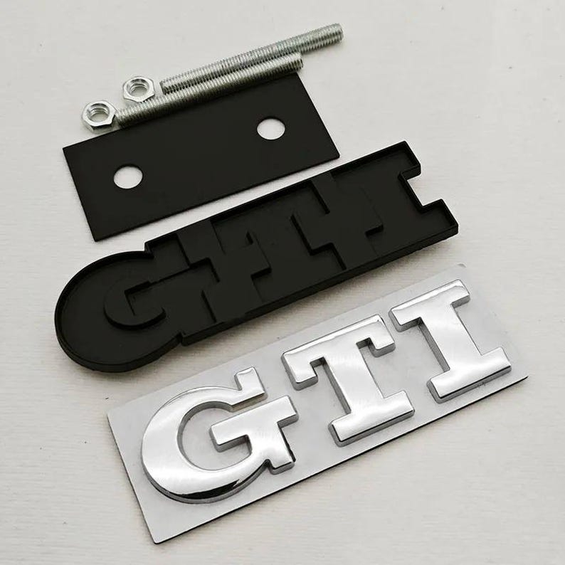 May include: A black and silver GTI emblem set, comprising a black base, silver letters, mounting hardware, and a black mounting plate. The GTI letters are in a bold, chrome-like font. Designed for automotive applications.