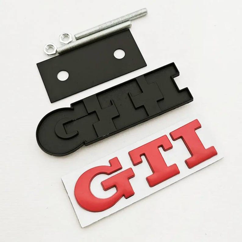 May include: A black GTI emblem with red GTI lettering, a black mounting bracket, and hardware. The emblem is a car accessory, likely for a vehicle's exterior. The parts are arranged on a white surface.