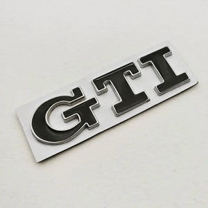 May include: A black and silver GTI badge. The letters are black with a silver outline, set on a rectangular silver backing. The badge is designed for automotive use.