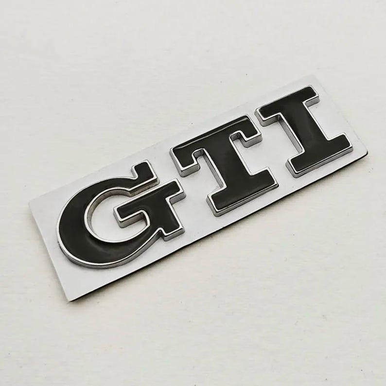 May include: A black and silver GTI badge. The letters are black with a silver outline, set on a rectangular silver backing. The badge is designed for automotive use.