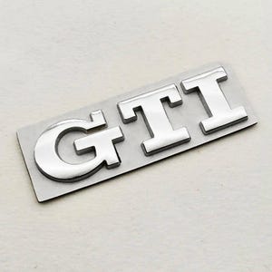 May include: A chrome GTI emblem, with the letters G, T, and I, on a rectangular silver backing. The letters have a reflective, metallic finish, suitable for automotive applications. The emblem is designed for cars.