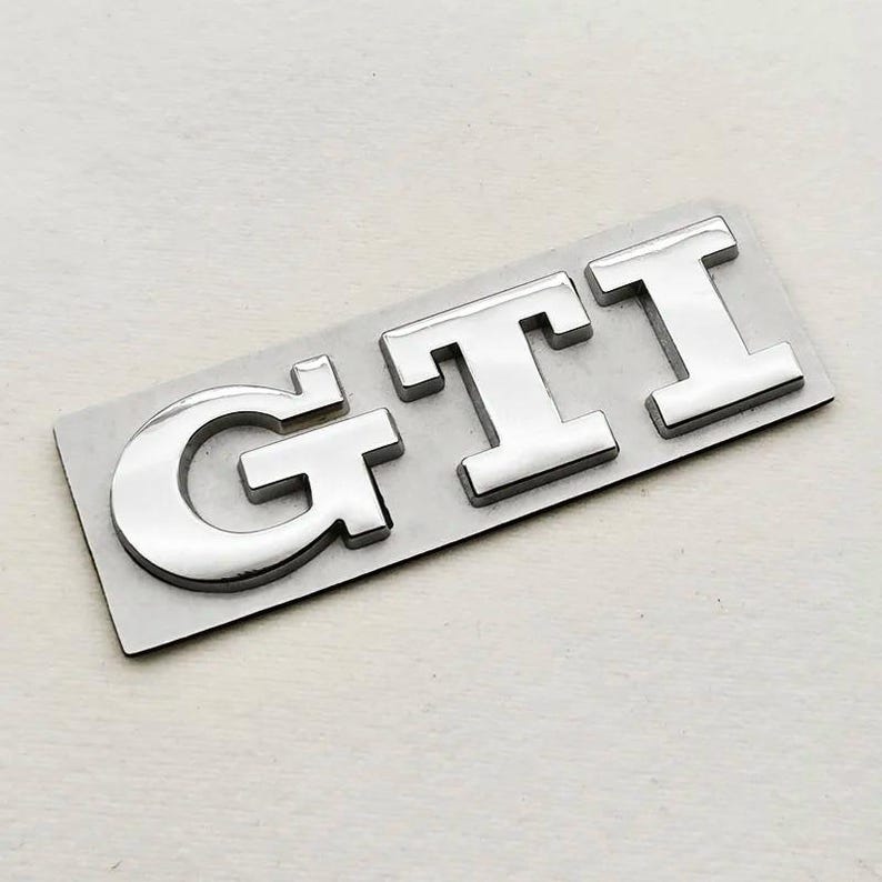 May include: A chrome GTI emblem, with the letters G, T, and I, on a rectangular silver backing. The letters have a reflective, metallic finish, suitable for automotive applications. The emblem is designed for cars.