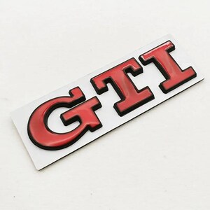 May include: A red and black GTI badge. The letters GTI are in a bold, blocky font with a glossy red finish and a black outline. The badge is rectangular and set against a white background.