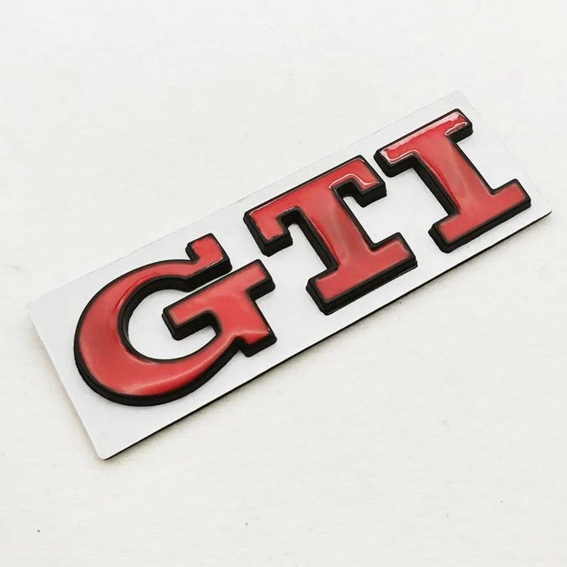 May include: A red and black GTI badge. The letters GTI are in a bold, blocky font with a glossy red finish and a black outline. The badge is rectangular and set against a white background.