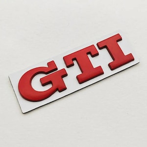 May include: Red GTI badge on a white rectangular backing. The letters are raised and in a bold font. The emblem is designed to be attached to a car, likely a Volkswagen GTI model.