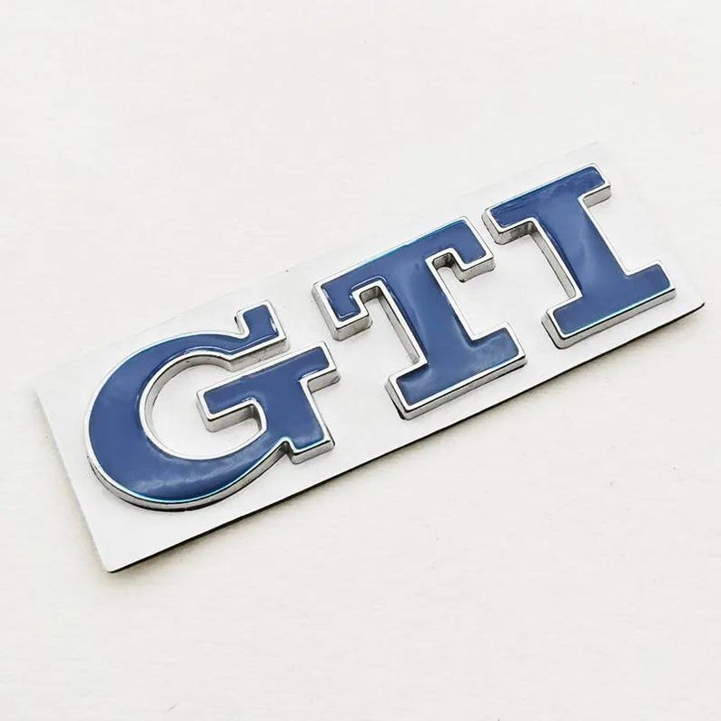 May include: A blue and silver GTI emblem. The letters G, T, and I are in a bold, sans-serif font. The letters are blue with a silver outline, mounted on a white rectangular base. The emblem is designed for automotive use.