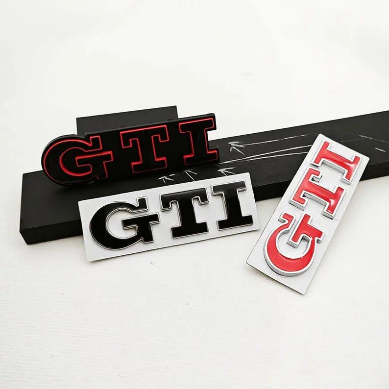 May include: Three GTI car emblems in black, red, and silver. The emblems are in various styles, including a black emblem with red trim, a black and silver emblem, and a red and silver emblem.