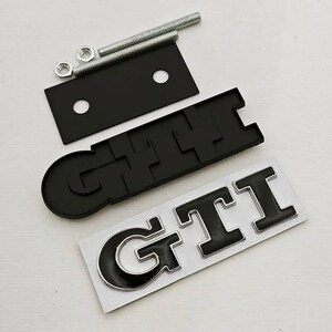 May include: Black GTI emblem with a silver outline, a black mounting plate, bolts, and nuts. The GTI emblem is on a white backing. These car parts are likely aftermarket accessories.