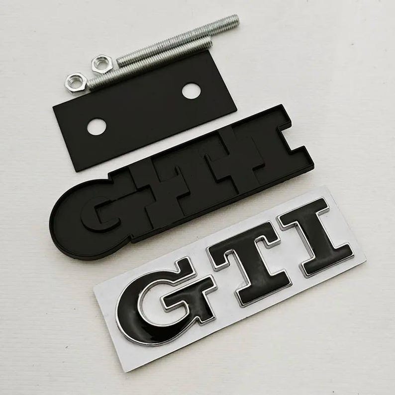 May include: Black GTI emblem with a silver outline, a black mounting plate, bolts, and nuts. The GTI emblem is on a white backing. These car parts are likely aftermarket accessories.