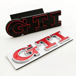 May include: Two GTI car emblems. One is black with red lettering, the other is red with a silver outline. The emblems are designed to be affixed to a vehicle. The GTI logo is a popular automotive design.