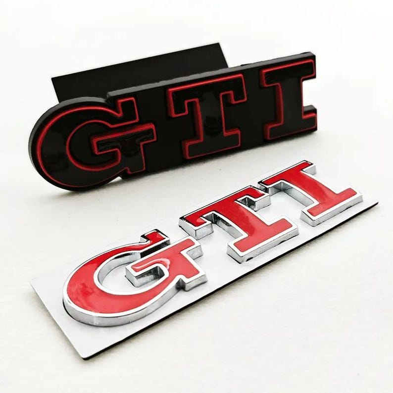 May include: Two GTI car emblems. One is black with red lettering, the other is red with a silver outline. The emblems are designed to be affixed to a vehicle. The GTI logo is a popular automotive design.