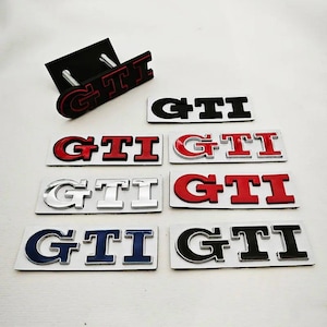 May include: A selection of GTI car badges in different colours, including red, black, silver, and blue. The badges are presented on a white surface, with one badge attached to a black mounting plate.