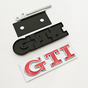 May include: A black GTI emblem with red lettering and a chrome outline, plus a black mounting plate, bolts, and nuts. The emblem is on a white surface. Automotive accessory.