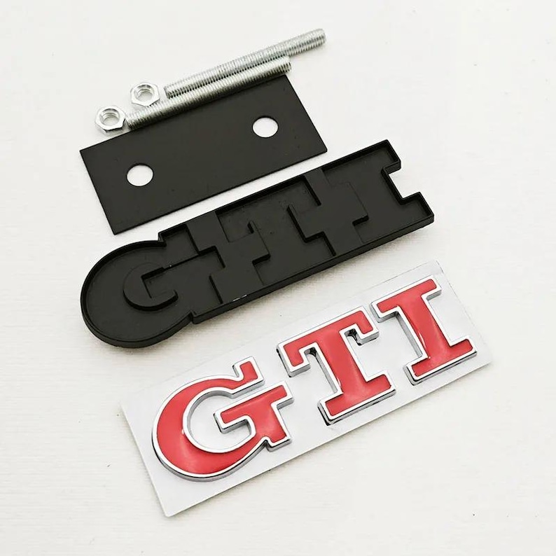 May include: A black GTI emblem with red lettering and a chrome outline, plus a black mounting plate, bolts, and nuts. The emblem is on a white surface. Automotive accessory.