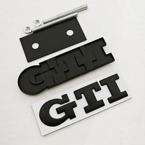 May include: Black GTI emblem set with mounting hardware. Includes a black rectangular plate with two holes, two bolts, two nuts, and two GTI emblems. The GTI emblems are black and have a raised design.