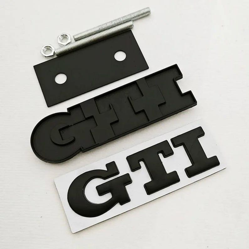 May include: Black GTI emblem set with mounting hardware. Includes a black rectangular plate with two holes, two bolts, two nuts, and two GTI emblems. The GTI emblems are black and have a raised design.