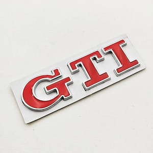 May include: A red and silver GTI badge. The letters "GTI" are in a bold, sans-serif font, with a red enamel finish and a silver outline. The badge is mounted on a rectangular silver backing.