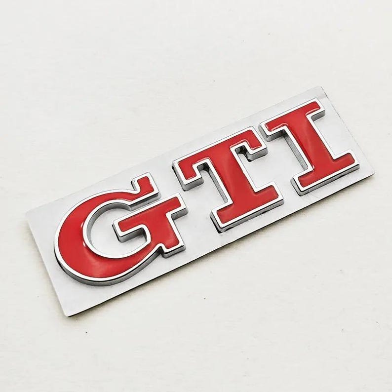 May include: A red and silver GTI badge. The letters "GTI" are in a bold, sans-serif font, with a red enamel finish and a silver outline. The badge is mounted on a rectangular silver backing.