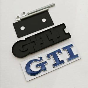 May include: A selection of car emblems and fixings. The image shows a black "GTI" badge, a silver and blue "GTI" badge, a black mounting plate, two bolts, and two nuts. The emblems have a shiny appearance.