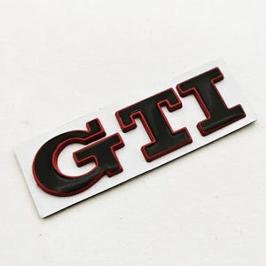 May include: Black and red GTI badge on a white background. The letters are black with a red border, giving a 3D appearance. This emblem is likely for a vehicle, possibly a Volkswagen.
