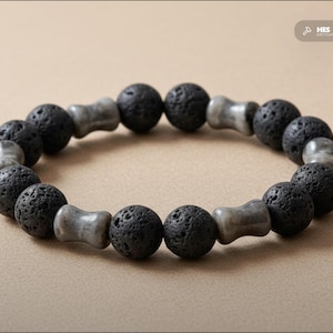 Könnte beinhalten: Armband mit abwechselnd schwarzen Lavasteinkugeln und grauen, zylindrischen Perlen. Das Armband wird vor einem neutralen Hintergrund präsentiert, der seine natürlichen Texturen und Farben hervorhebt. Der Umfang beträgt ca. 18 cm.