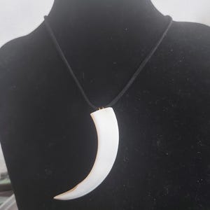 Pode incluir: Um pendente branco em forma de dente curvo suspenso por um cordão preto fino. O pendente tem uma superfície lisa e sólida que se estreita para uma ponta na base. Está preso ao cordão por uma pequena argola de metal na cor dourada.