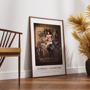 Puede incluir: Un cuadro enmarcado con una mujer con un vestido, con el texto "FLORENCE + THE MACHINE EVERYBODY SCREAM" en la parte inferior. El cuadro está apoyado contra una pared blanca, junto a una silla de madera y plantas secas decorativas.