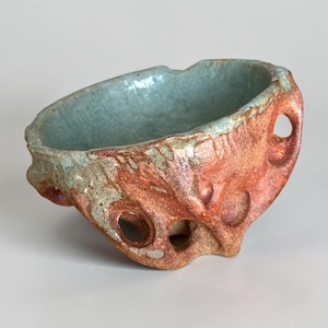 May include: A small, hand-formed ceramic bowl with a textured, reddish-brown exterior featuring several circular cutouts. The interior of the bowl is glazed in a smooth, pale blue-green finish that contrasts with the rough, earthy outer surface.