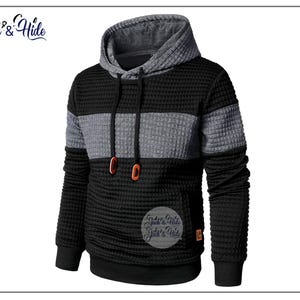 Op de afbeelding: Zwart en grijze hoodie met een textuur van wafelbreisel. De hoodie heeft een grijze baan over de borst en een grijze capuchon. Het heeft een kangoeroezak en oranje oogjes. De merknaam "Jack's & Hide" is zichtbaar.