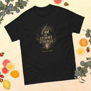 May include: Black t-shirt with the text "I AM THE STORM'S STILLNESS" in gold lettering. The design includes an abstract, swirling graphic. The brand name "Pelley's Studio" is printed below the text.