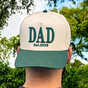 May include: A cream-colored baseball cap with a dark green brim and dark green embroidered text on the front that reads DAD Est.2025. The cap features a structured crown with a button on top and a flat brim design in a two-tone color scheme.