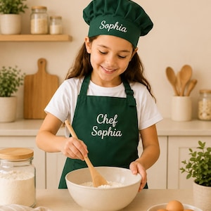 May include: A dark green chef hat and matching apron set featuring white embroidered text. The hat displays the name Sophia, while the apron is embroidered with the words Chef Sophia in a script font. The set is made of solid dark green fabric.