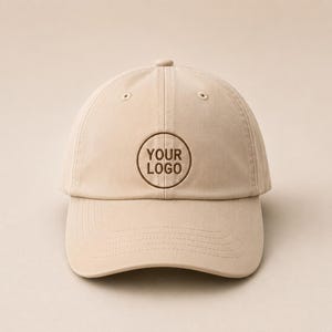 May include: A beige baseball cap featuring a circular brown embroidered design on the front panel. The embroidery consists of a thin circular outline with the text YOUR LOGO centered inside. The cap has a curved brim and stitched eyelets.