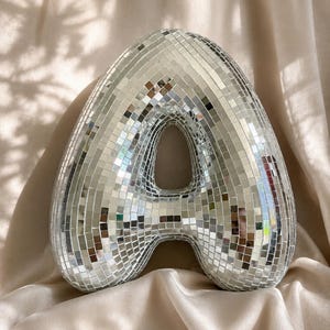 May include: A three-dimensional letter A covered in small, square, reflective silver mirror tiles. The surface of the letter is composed of many individual glass facets that create a mosaic effect, reflecting light across the curved, rounded shape.