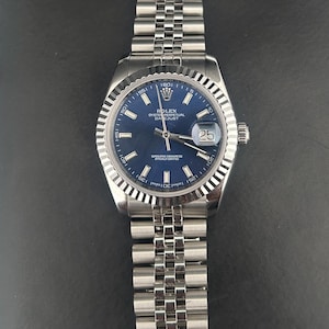 Puede incluir: Un reloj Rolex Oyster Perpetual Datejust de acero inoxidable con esfera azul y bisel plateado texturizado. El reloj tiene un brazalete de eslabones plateado. La fecha se muestra a las 3 en punto.