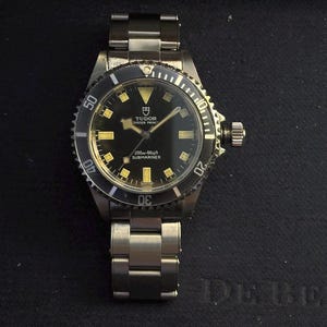 May include: A black and silver Tudor Oyster Prince Submariner watch. The watch has a black dial with gold-colored hour markers and hands. The bezel is black with white markings. The watch band is silver and made of metal.