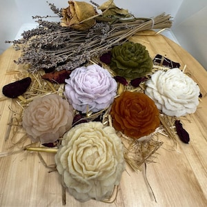 Peut inclure: Un assortiment de savons faits à la main en forme de fleurs de différentes couleurs, dont lavande, vert et marron, disposés sur une surface en bois avec de la lavande séchée et de la paille.