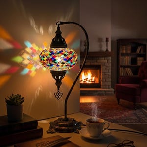 Puede incluir: Una lámpara de mesa decorativa con una pantalla de mosaico de vidrio multicolor. La lámpara tiene un marco de metal oscuro curvado y una base detallada. Está sobre una mesa con libros, una taza de té y gafas.